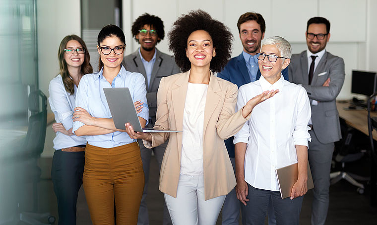 Eine Gruppe bestehend aus Frauen und Männern, die sich im Büro befinden. Dir Frau in der Mitte hält einen Laptop in der Hand. 